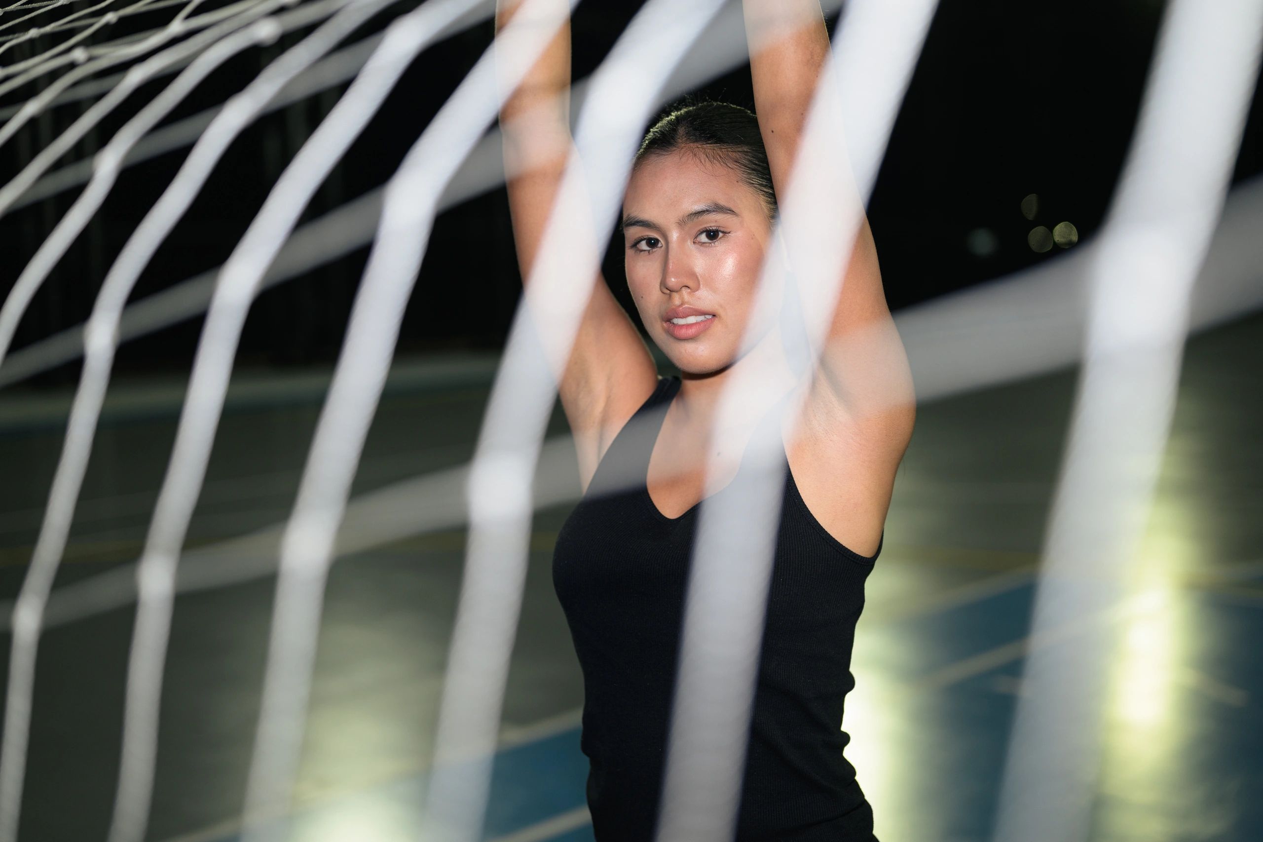 Athletic Asian Thai woman looking at camera with arms raised behind soccer goal net on outdoor court at night
